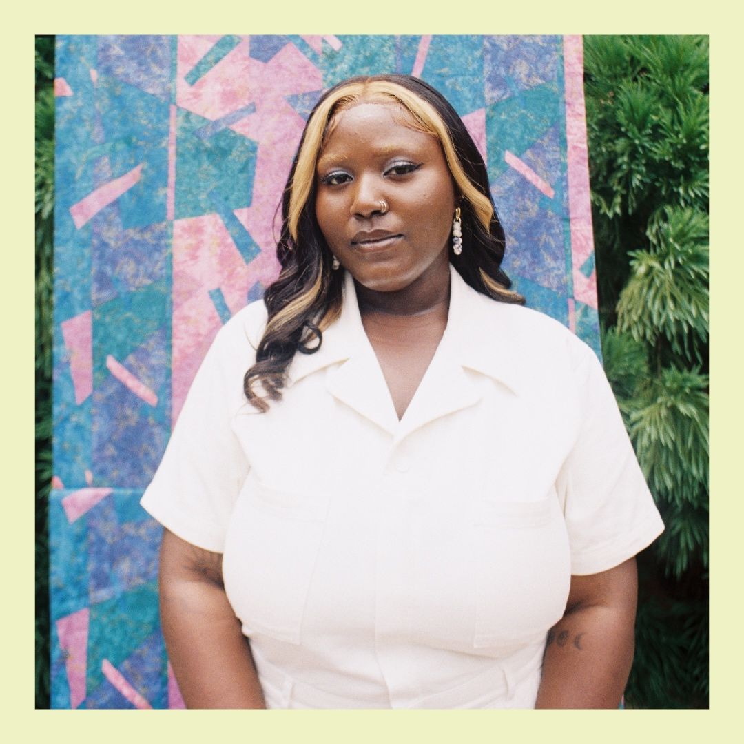 Black woman with brown and blonde hair stands in front of pink and blue backdrop